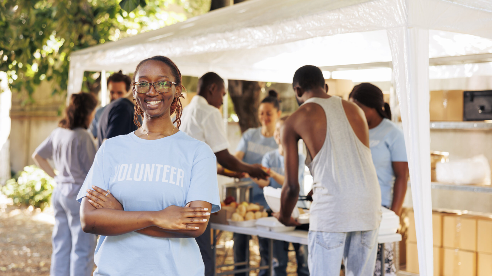 Young black woman with glasses stands outdoors, arms crossed, looking at the camera. Diverse group of volunteers supports a non-profit program dedicated to hunger relief and helping needy individuals.