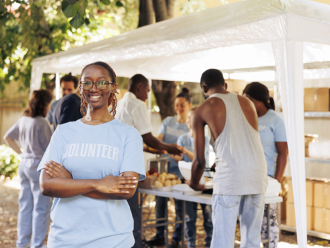 Young black woman with glasses stands outdoors, arms crossed, looking at the camera. Diverse group of volunteers supports a non-profit program dedicated to hunger relief and helping needy individuals.